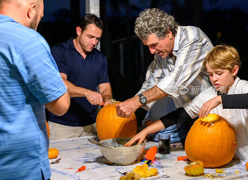 Two fathers and grandparent carve pumpkins together with their children